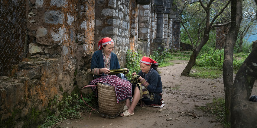 Two women dressed in traditional clothes are sitting outside an old building with a basket.