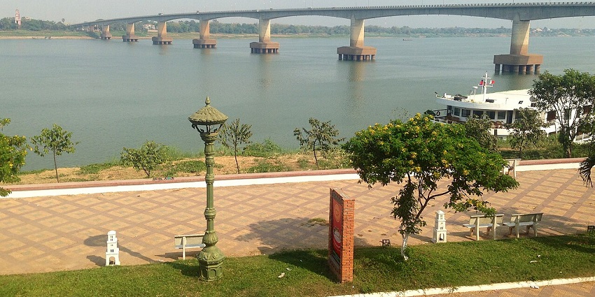 Long concrete bridge spanning across a wide river with riverside trees, and a docked boat.