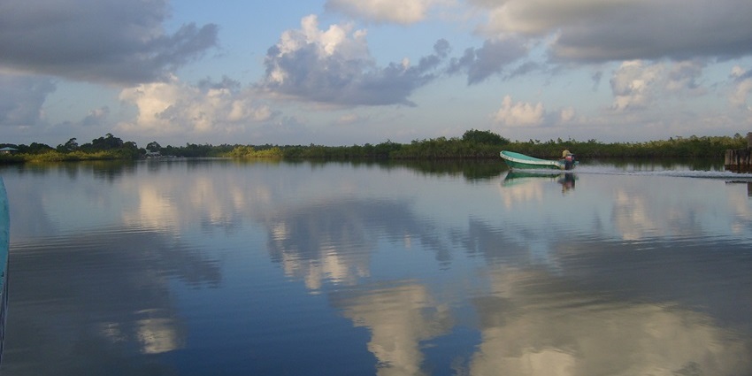 Placencia Lagoon in Belize with a man on the green motorboat and reflections of clouds, one of the best things to see in Placencia Belize.