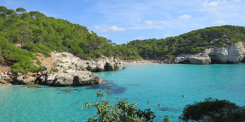 Turquoise Bay is surrounded by rocky cliffs and lush green forest under a bright blue sky.