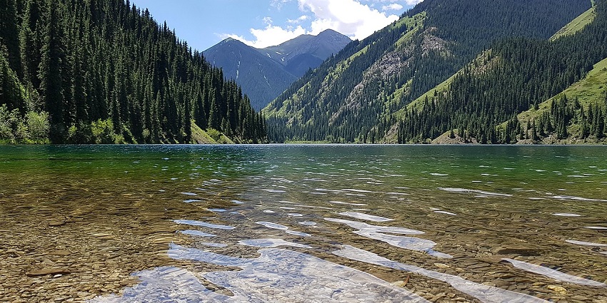 Kolsai Lake with calm, turquoise water, surrounded by pine-covered slopes and rocky hills.