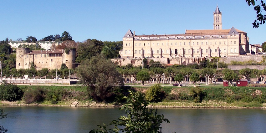 River view with leafy trees, rustic rooftops, and a suspension bridge, amongst the best places to visit near Bordeaux.