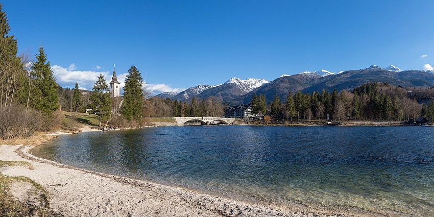 Tranquil lake with pebbled shore, stone bridge, and church tower at a breathtaking places to visit near Ljubljana.