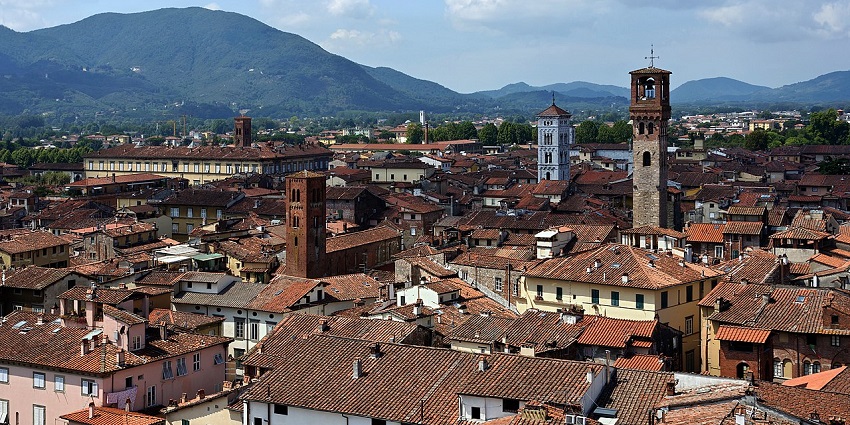 Historic town with medieval towers, red-tiled roofs, and rolling hills in the background.
