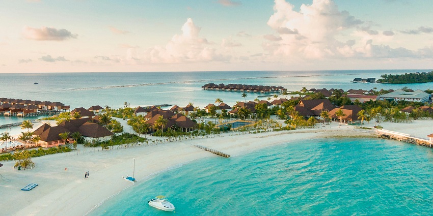 The Maldives with distant buildings of the resorts, boats near the sandy shore and green palm trees.
