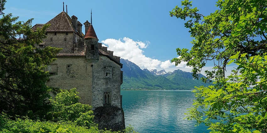 Medieval stone castle by a serene lake, framed by lush greenery and distant mountain peaks, one of the places to visit near Geneva.