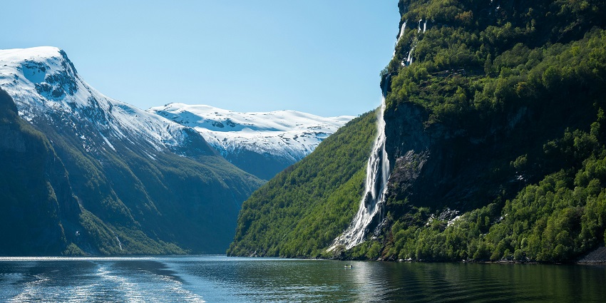 A waterfall flowing from a cliff covered in greenery into a water body and distant snow mountains.