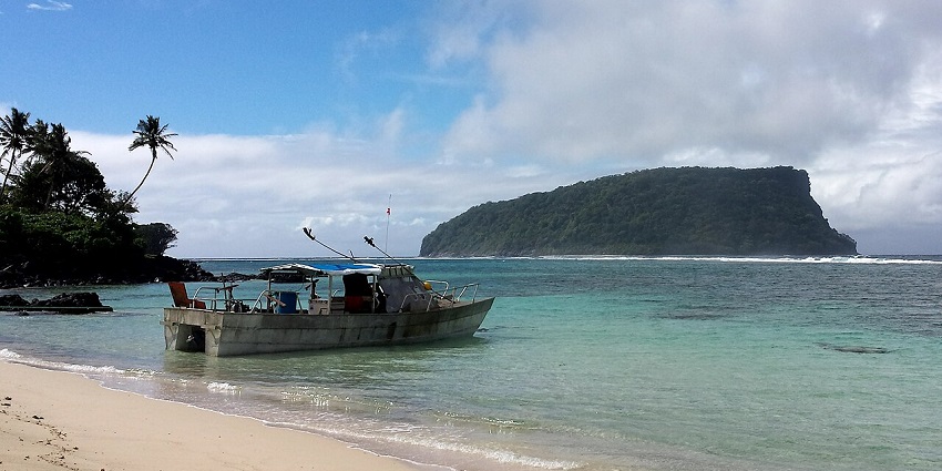 A small fishing boat rests on the shore of a tropical beach with a lush island rising in the distance.