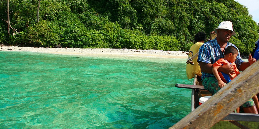A family enjoys a boat ride in the clear turquoise waters near a lush green island shore.