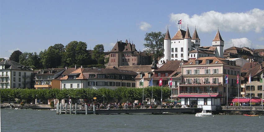Scenic lakeside view with a white castle, historic buildings, and flags under a blue sky.