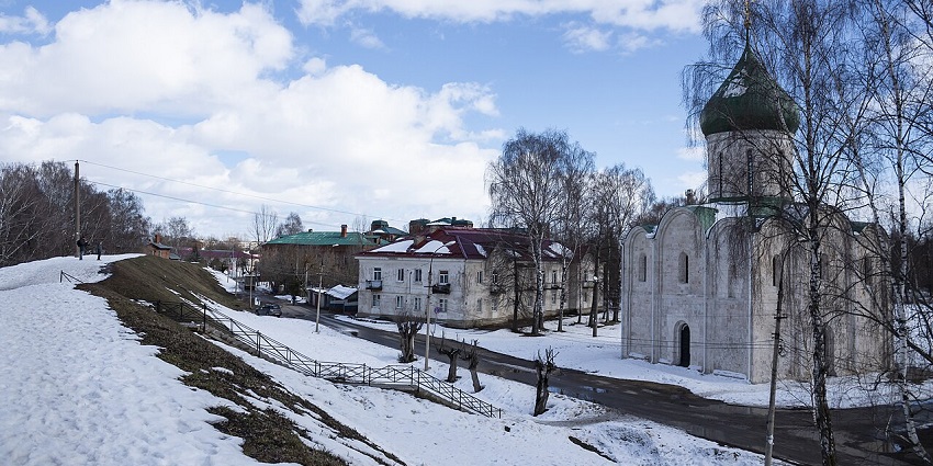 Snowy landscape with a Russian Orthodox church and nearby village houses under blue sky.