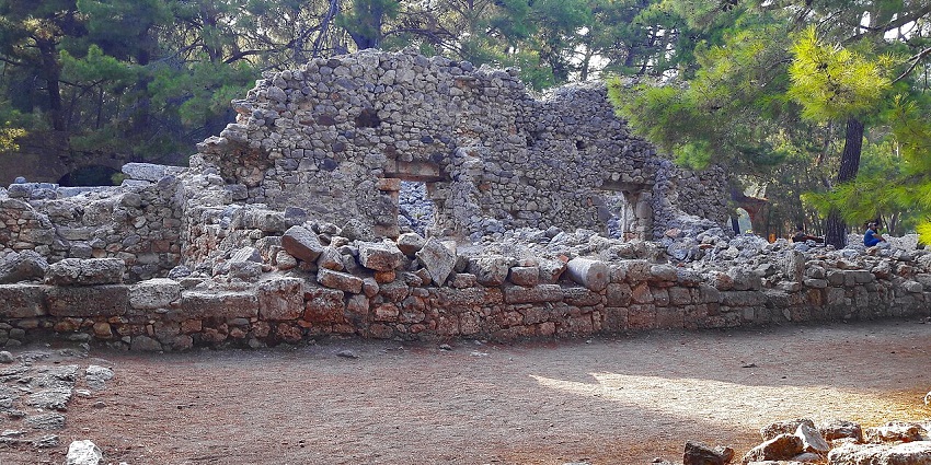 Ancient stone ruins at Phaselis are surrounded by dry earth and pine trees in the background.