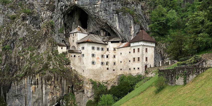 Dramatic stone castle nestled in a cliffside cave, set against lush green hills.