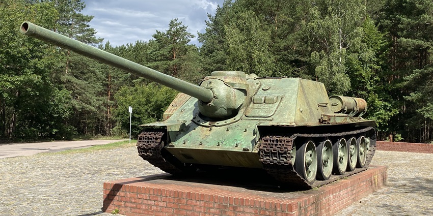 A preserved Soviet tank is displayed on a brick pedestal surrounded by dense forest trees, one of the places to visit near Berlin.
