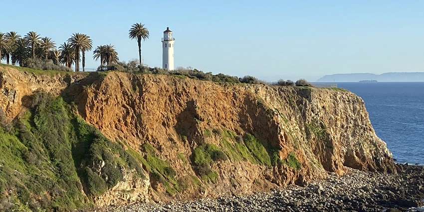 Scenic coastal view with a white lighthouse, cottages, grassy dunes, and sea under a cloudy sky.