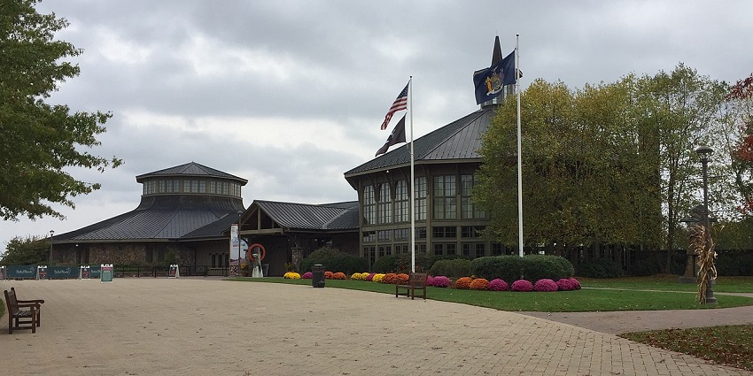 Octagonal-roofed building with flags, trees, and colourful flower beds under a cloudy sky.