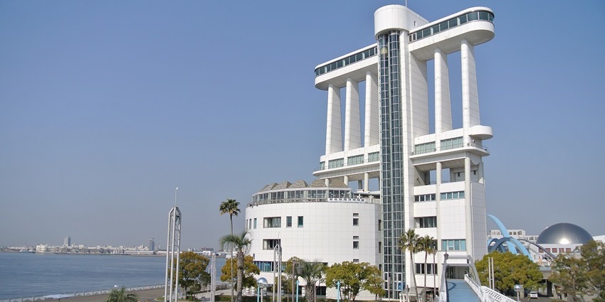 Futuristic white building with tall pillars beside a waterfront, surrounded by trees.