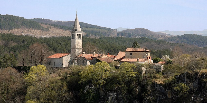 Hilltop village with terracotta rooftops and a stone church tower, surrounded by dense forest.