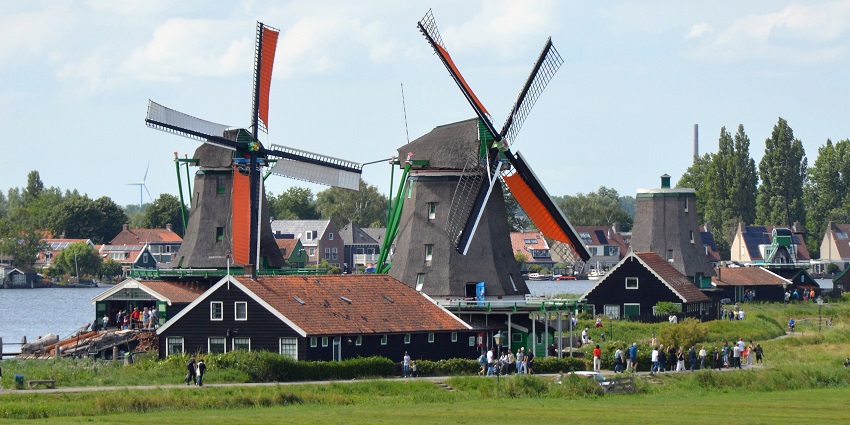 Historic windmills by a calm river, with green banks and a cloudy sky in the background.