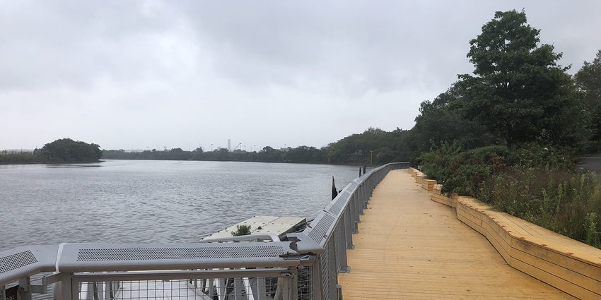 Wooden boardwalk next to a calm river under clear sky with distant trees in the background.