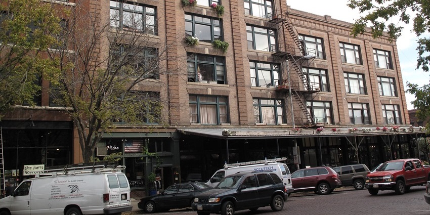 Brick building with large windows, fire escape stairs, and parked cars lining the street.