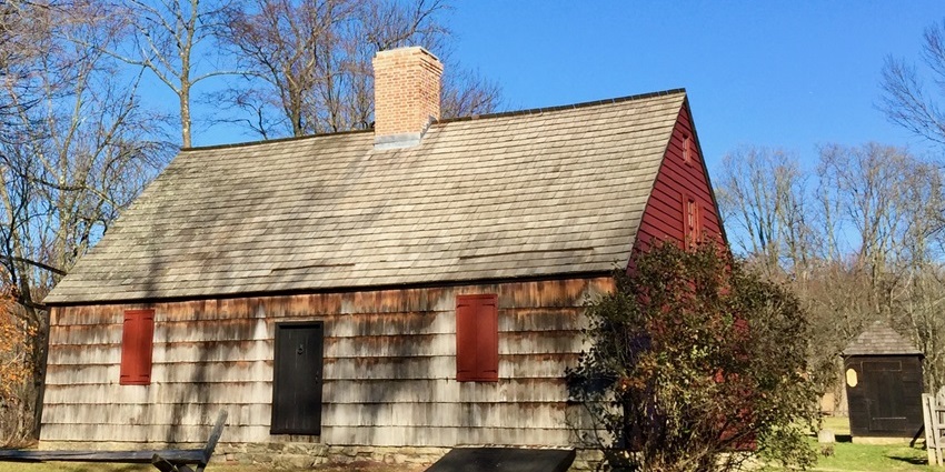 Small historic wooden house with red shutters, a stone foundation, and a brick chimney.