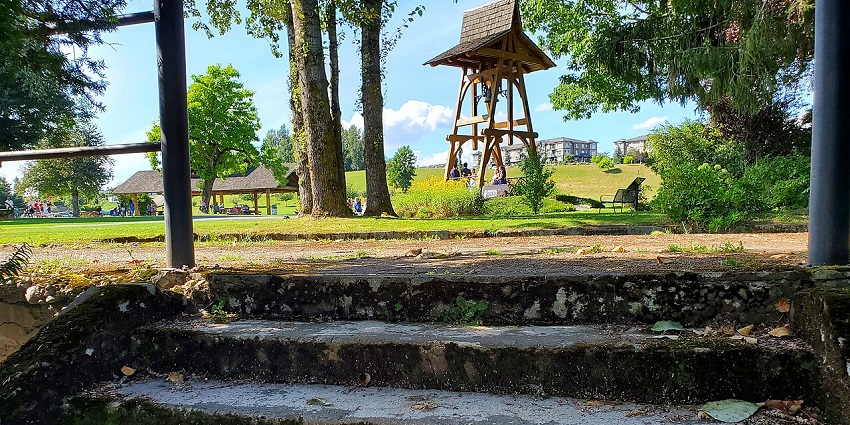 Mossy stone steps lead to a sunny park with a wooden bell tower, trees, and people gathering.