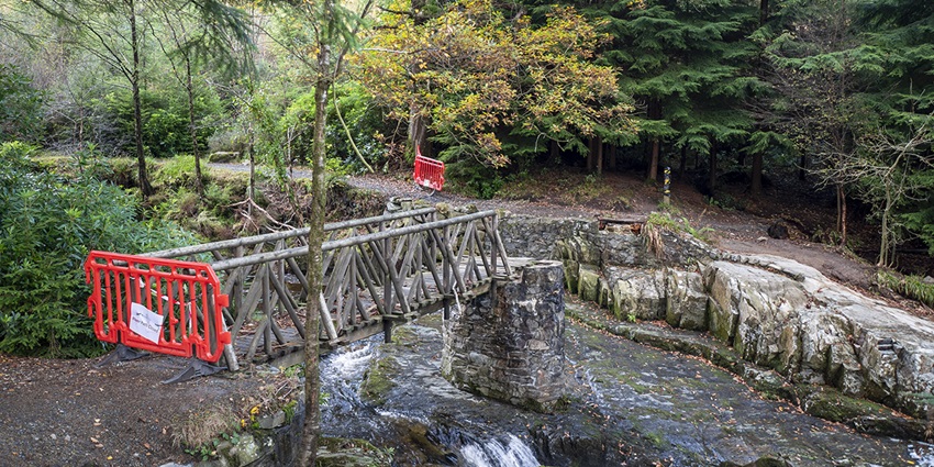 Wooden footbridge over a rocky stream in a forest, closed off with red safety barriers.