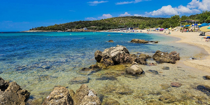 Golden sand and rocky shoreline at Gidaki Beach with clear blue waters in the background.