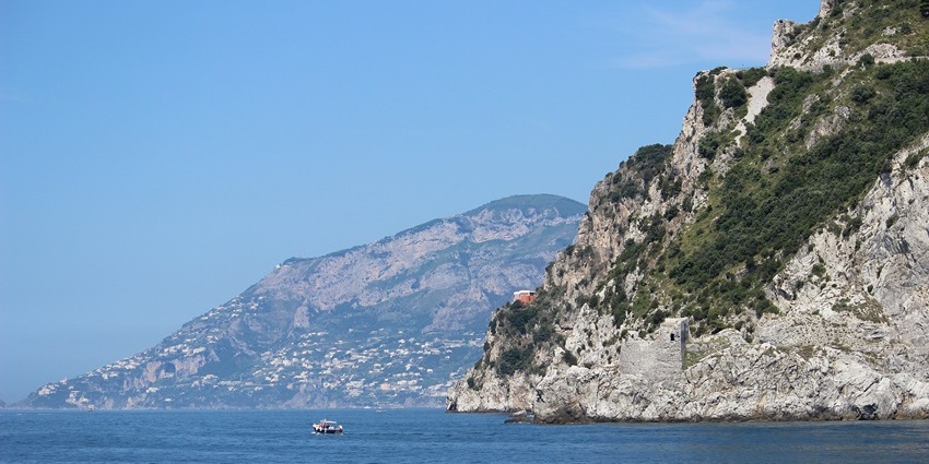 Amalfi Coast cliffs with terraced homes and a small boat floating on the calm blue sea.
