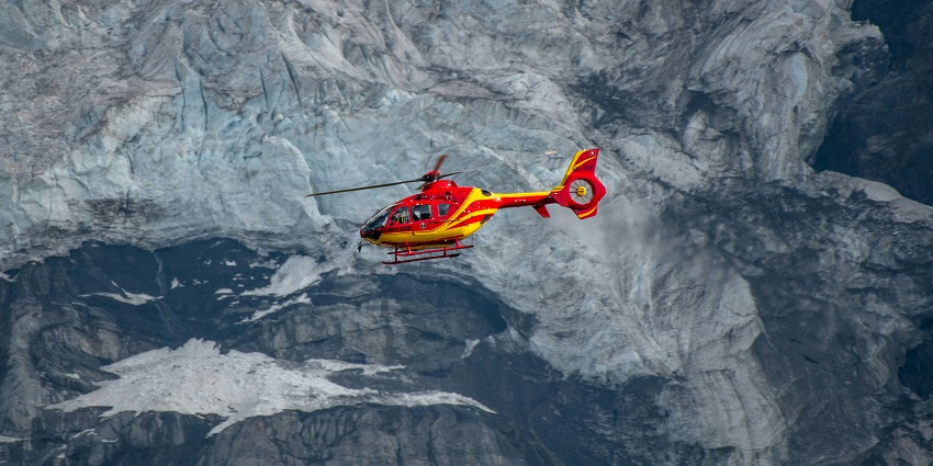 Helicopter flying over a glacier with snowy mountains and icy terrain below.