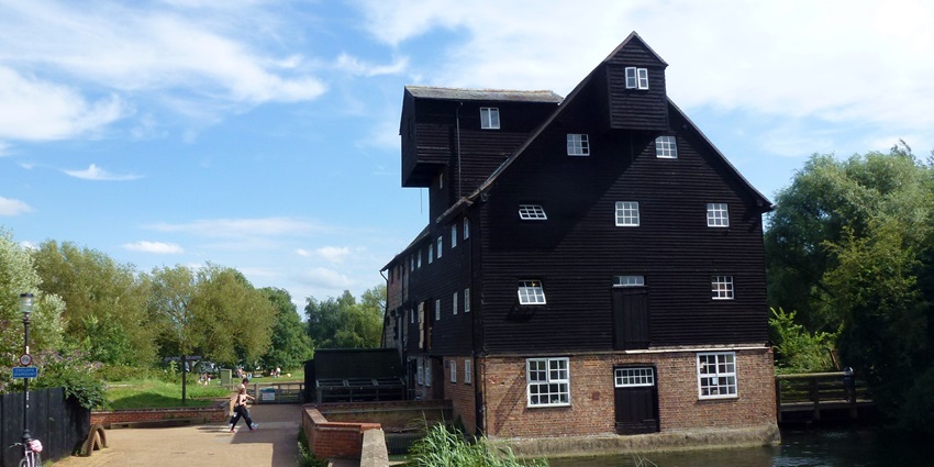 Houghton Mill, a black timber watermill beside a river with swans, trees, and blue sky.