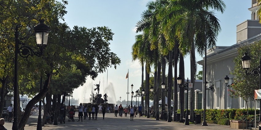 Tree-lined walkway with people strolling toward a large fountain and sculptures, one of the best things to see in Old San Juan.