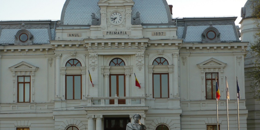 Elegant 19th-century city hall with arched windows, Romanian flags, and a statue in front.