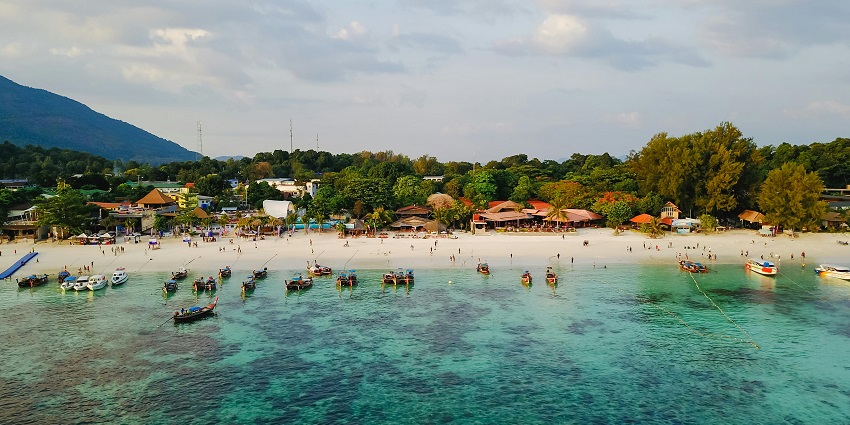 A beach in Thailand with clear blue water, boats, and a sandy shore, places to visit in February near India.
