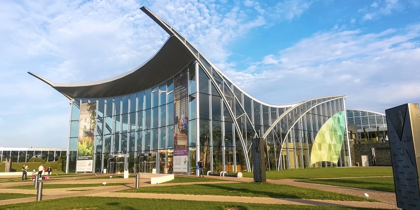 Modern glass building with sweeping curved roof design set against a blue sky with clouds.