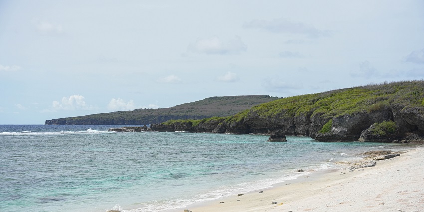 Wide sandy beach on Tinian Island with clear blue waters and gentle waves.