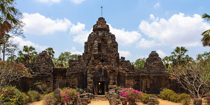 Ancient stone temple with weathered architecture, flowers, and a bright blue sky above.