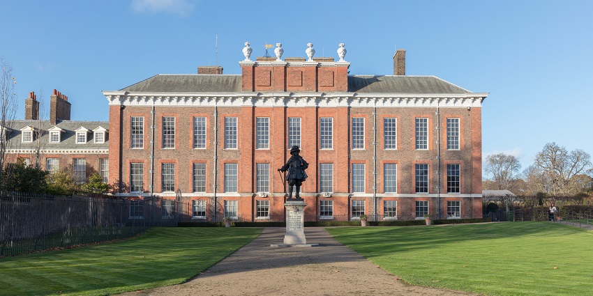 Front view of Kensington Palace with red brick exterior and a black statue in front.