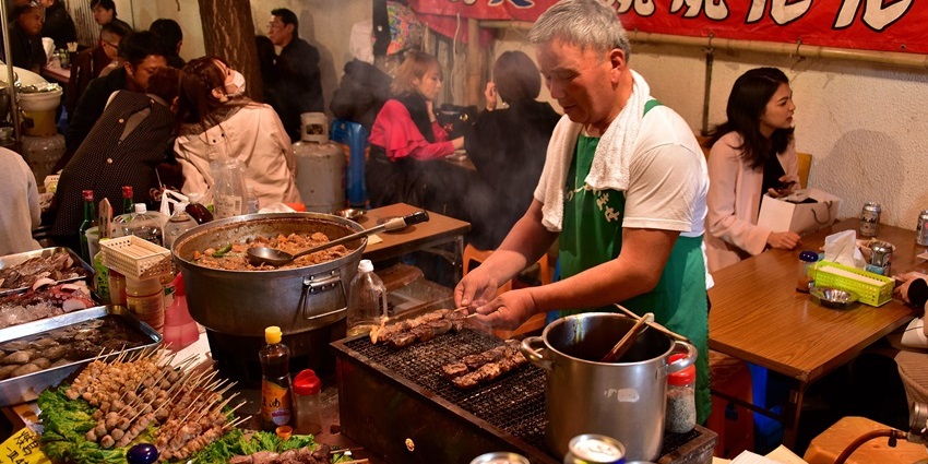 A street food vendor grills skewers for customers at a crowded Tokyo night market.