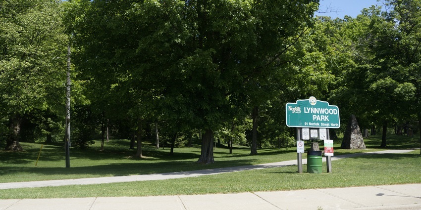Open grassy area at Lynnwood Park in Simcoe, with trees and a wooden entrance sign.