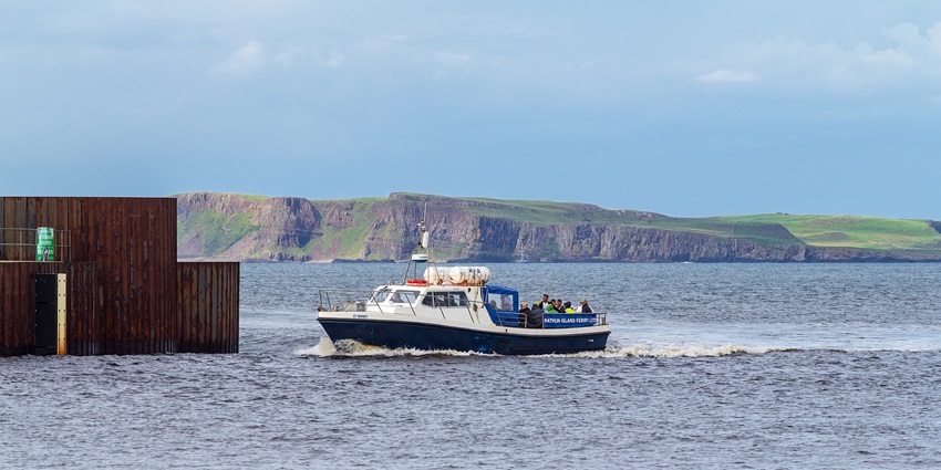 A small passenger ferry sailing past a wooden pier with cliffs and hills in the background.