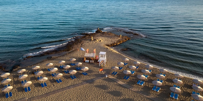 Aerial view of a sandy beach with straw umbrellas, wooden sunbeds, and calm blue sea.
