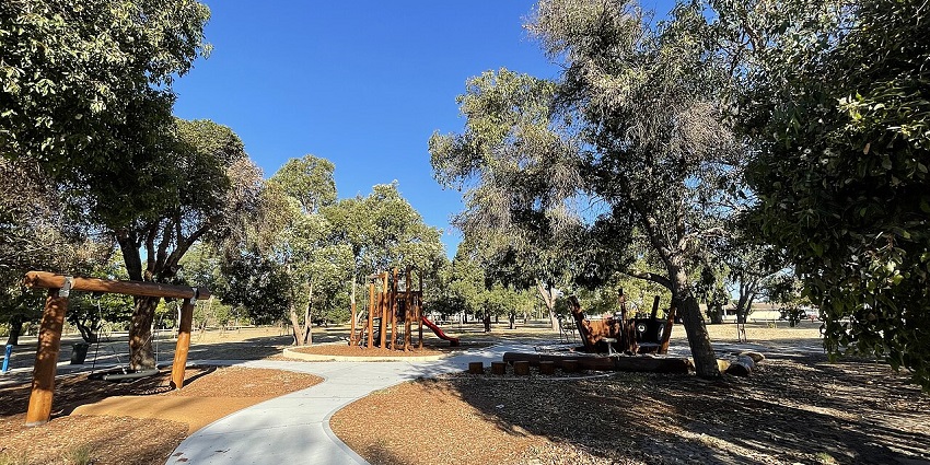 Sunny park with trees, walking path, wooden playground, and a pirate ship play structure.