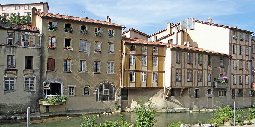 -Riverside buildings with rustic facades and flower-filled balconies on a sunny afternoon.