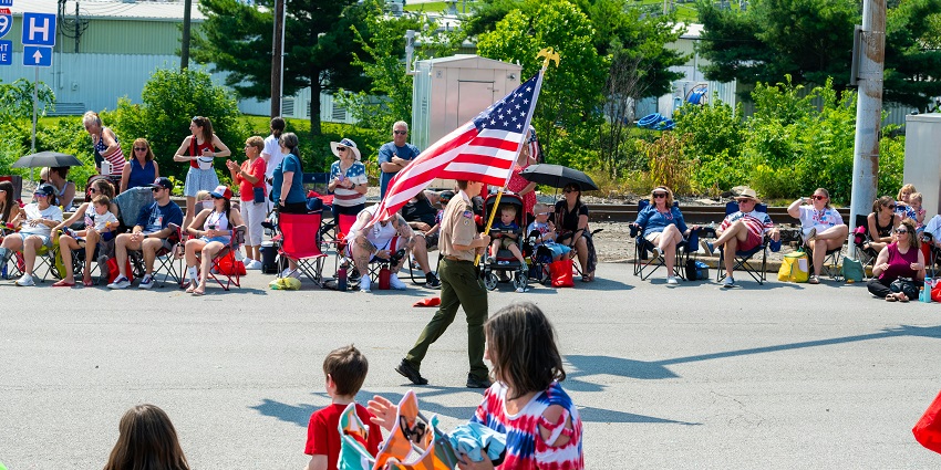 A parade where a Boy Scout is walking the flag of the USA while onlookers are cheering.