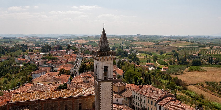 A stone bell tower with a pointed spire rising above a village, surrounded by countryside views.
