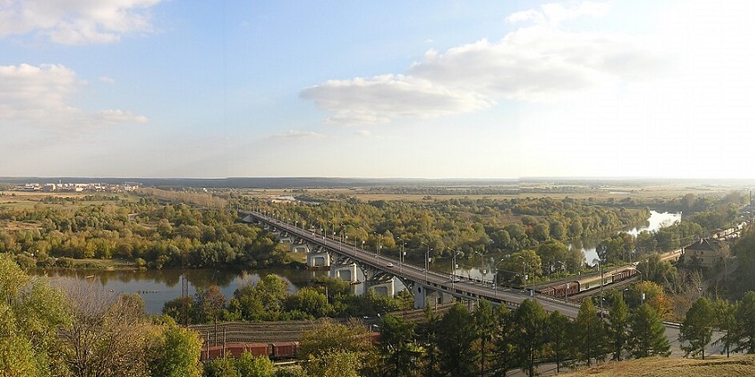 Panoramic view of a bridge over a river surrounded by dense greenery and open countryside.