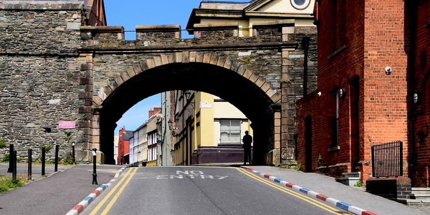 People stroll by a brick building and old stone wall on a quiet, slightly inclined street, one of the best things to see in Northern Ireland.