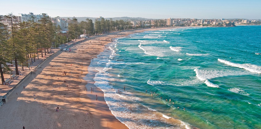 Aerial view of a beach with turquoise waves, golden sand, and a city in the distance.
